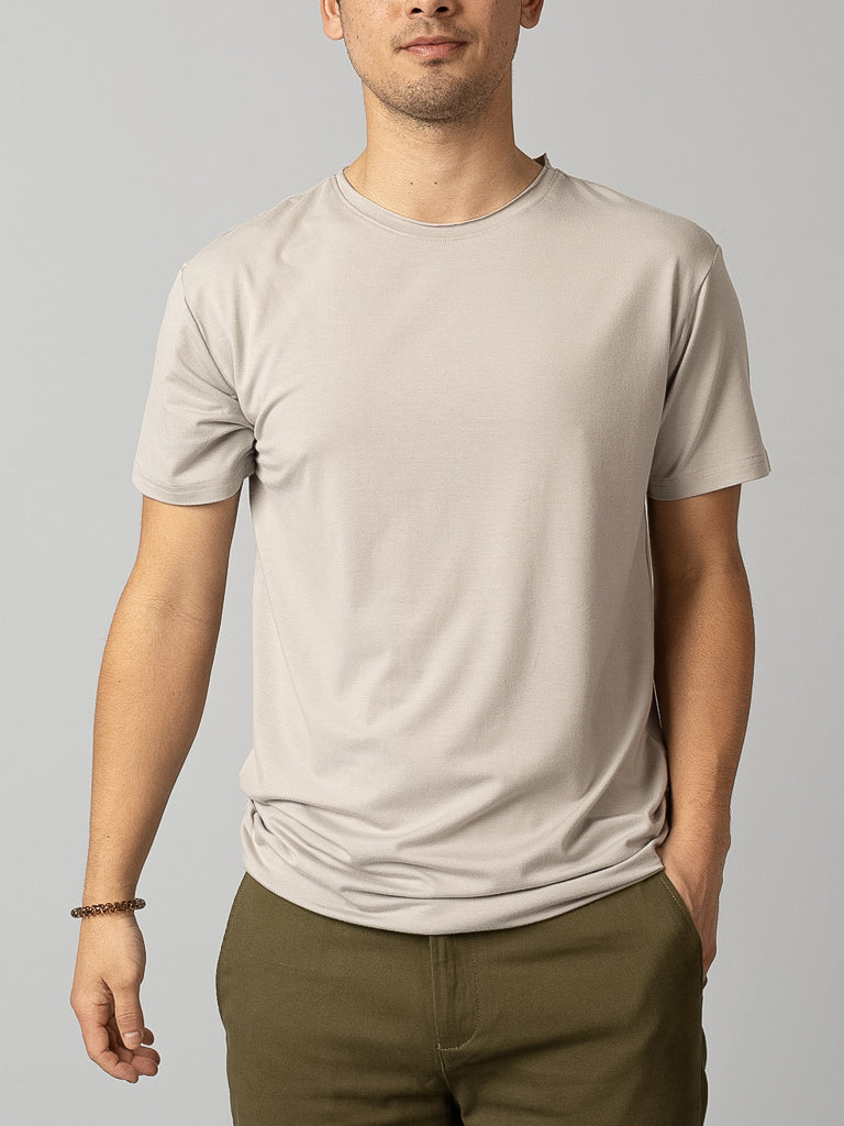 A person in a plain light gray Bello Bamboo Blend Rolled Neck Tee from ID Menswear and olive-green pants stands against a neutral background. One hand is in their pocket, the other by their side, embodying sustainable clothing. Their head is cropped out of the image.