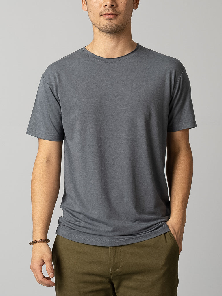 A person wearing an ID Menswear Bello Bamboo Blend Rolled Neck Tee in gray, paired with olive green pants, stands against a neutral background. One hand is in a pocket, the other rests by their side. They also wear a brown beaded bracelet, showcasing sustainable fashion with understated elegance.