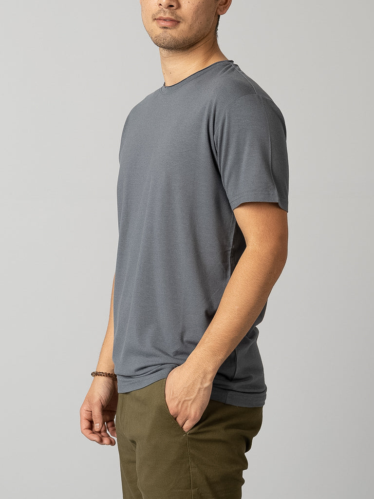 A person in a Bello Bamboo Blend Rolled Neck Tee from ID Menswear and olive-green pants stands against a gray background. The sustainable attire complements their relaxed pose as they face right, with their left hand casually in their pocket.