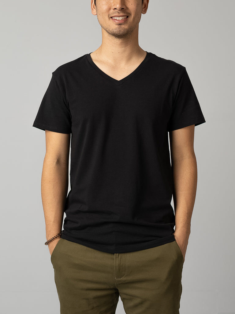 A person in an ID Menswear Form V Neck Tee, styled in a form-fitting black design, pairs it with olive-green pants. They stand against a plain gray background, hands casually in pockets and wearing a neutral expression. The image is cropped from shoulders to thighs, perfect for a casual snapshot.