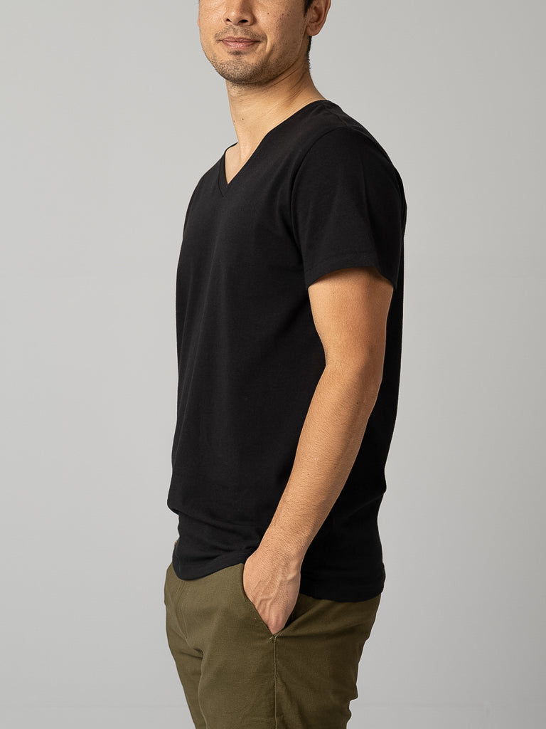 A man wears an ID Menswear Form V Neck Tee in black and green pants, standing confidently with his left hand in his pocket against a gray backdrop, perfect for casual occasions.