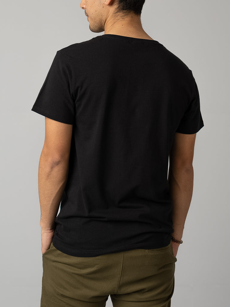 A person in ID Menswears form-fitting Form V Neck Tee and olive green pants stands with their back to the camera, hands in pockets against a neutral gray backdrop, ideal for casual occasions.