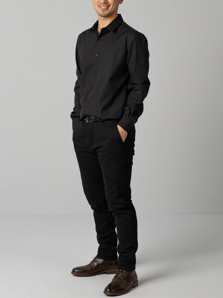 A person poses casually with hands in pockets, wearing an ID Cotton Dress Shirt from ID Menswear in slim-fit black, matched with pants and brown shoes. The plain light gray background accentuates the outfits casual yet smart appeal.