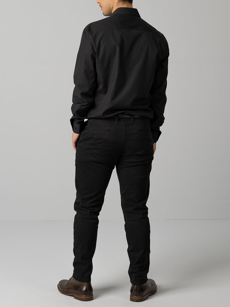 A man in an ID Menswear slim fit black shirt, with black pants and brown shoes, stands with his back to the camera against a plain gray background.