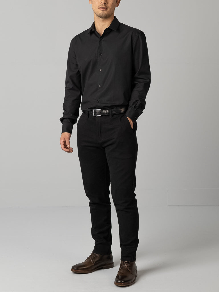 Standing against a neutral backdrop, someone is wearing an ID Cotton Dress Shirt in black slim fit from ID Menswear with black pants, hands in pockets. A sleek black belt and brown shoes complete the formal but monochromatic look.