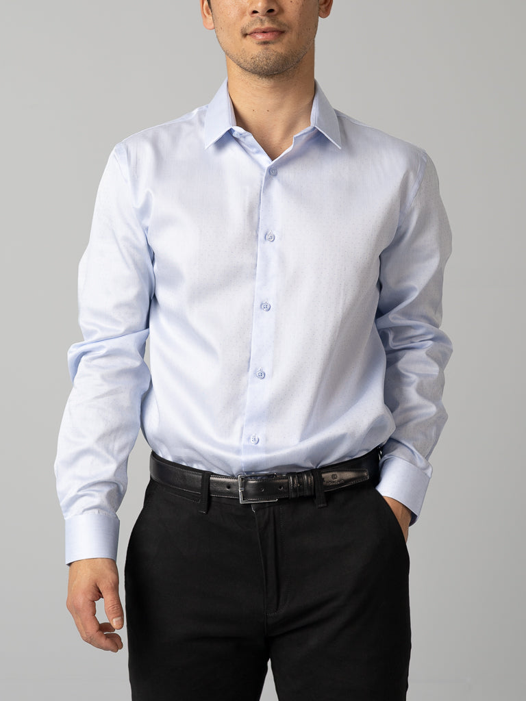 A man in an ID Menswear slim fit light blue cotton dress shirt, accompanied by black trousers and a black belt, stands against a gray background with his hands at his sides.