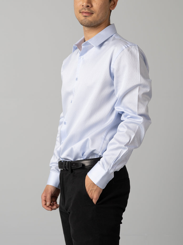 A person stands in a relaxed pose wearing an ID Menswear ID Cotton Dress Shirt in Slim Fit, light blue, paired with black pants and a black belt against a plain gray background.
