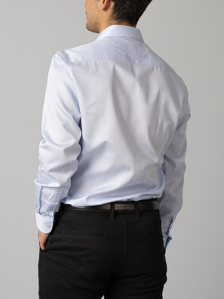 With their back to the camera, a person wears an ID Menswear light blue ID Cotton Dress Shirt in slim fit and black pants with a black belt, right hand casually in their pocket against a plain light gray background.