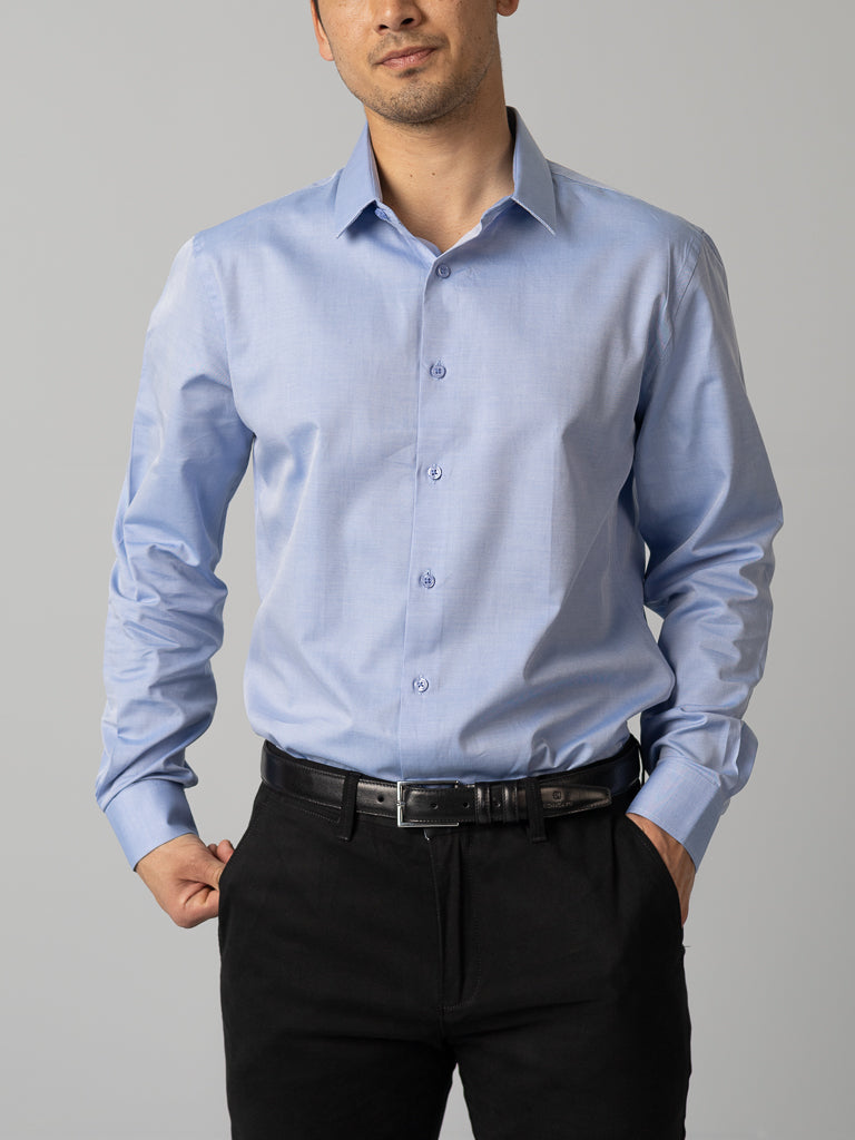 A man in an ID Menswear light blue Cotton Dress Shirt (slim fit) and black pants with a belt stands against a plain background, one hand in his pocket.