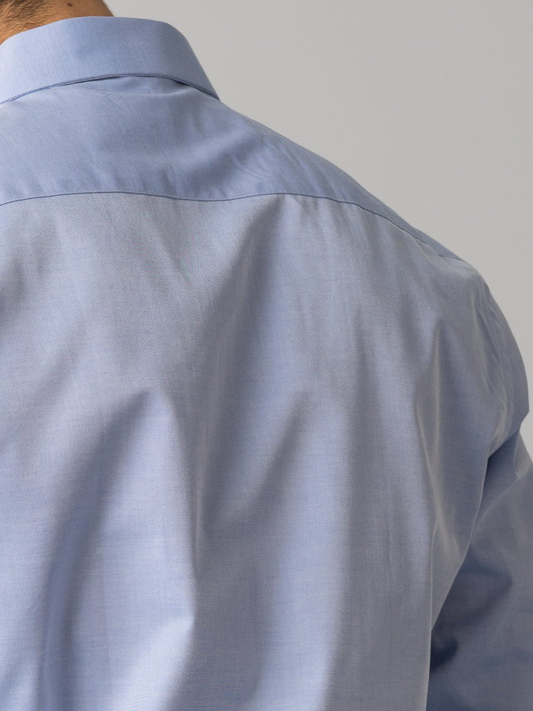 A close-up displays the back of an individual wearing an ID Menswear, light blue ID Cotton Dress Shirt in Slim Fit. Slight wrinkles appear on the fabric with visible collar and shoulder seam against a plain background.
