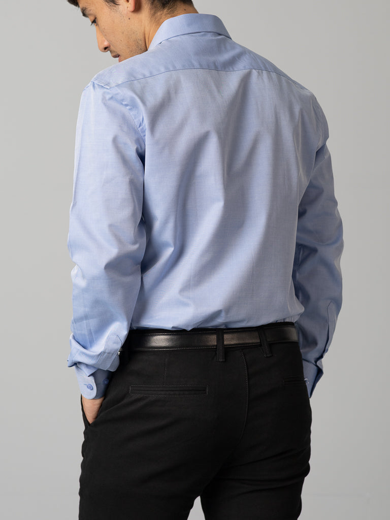 A person with short dark hair is seen from behind wearing an ID Menswear ID Cotton Dress Shirt in Slim Fit, light blue and neatly tucked into black pants with a sleek black belt. They stand against a plain gray background.
