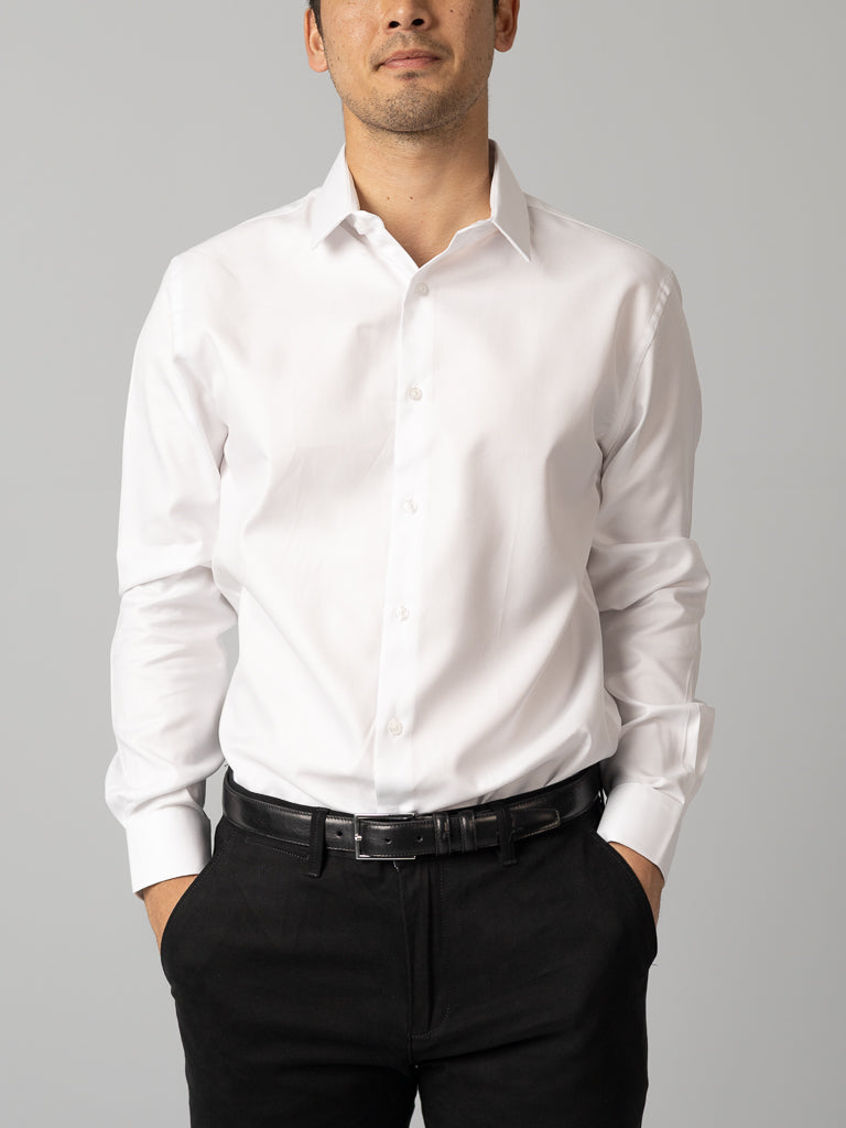A man stands against a plain light gray background, wearing the ID Menswear Cotton Dress Shirt in slim fit and black pants with a black belt. His hands are in his pockets, and his face is partially visible.