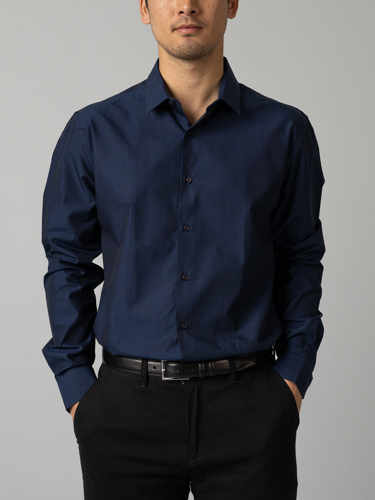 A man wears an ID Menswear Cotton Dress Shirt in Slim Fit, dark blue, paired with black pants, standing with hands in pockets against a plain light gray background.
