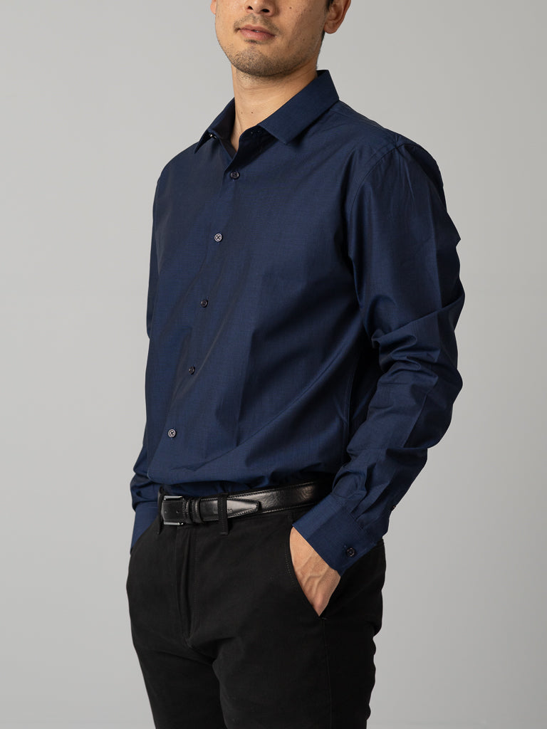 A man models the ID Menswear Cotton Dress Shirt in slim fit (dark blue) with black pants, standing sideways with one hand in his pocket against a plain light gray background.
