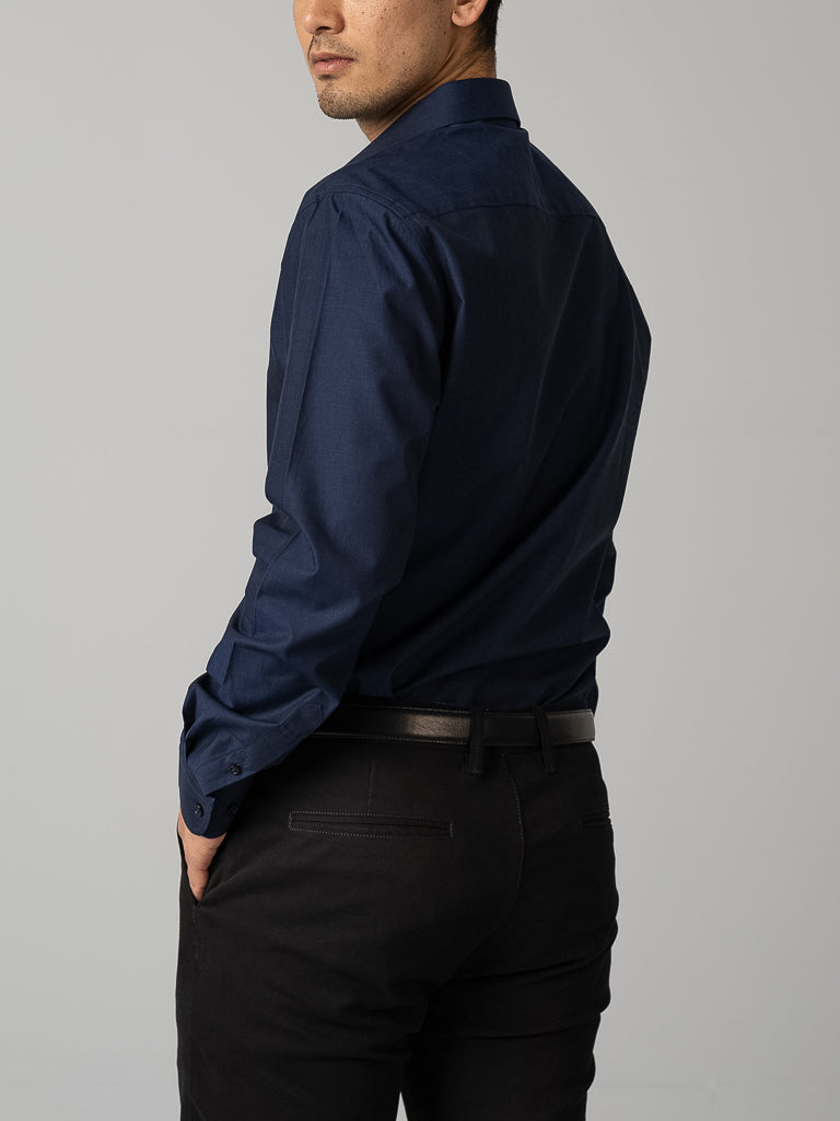 A man in an ID Menswear Cotton Dress Shirt, slim fit, dark blue, with black trousers stands against a plain light gray background, back turned and glancing over his shoulder, left hand in his trouser pocket.