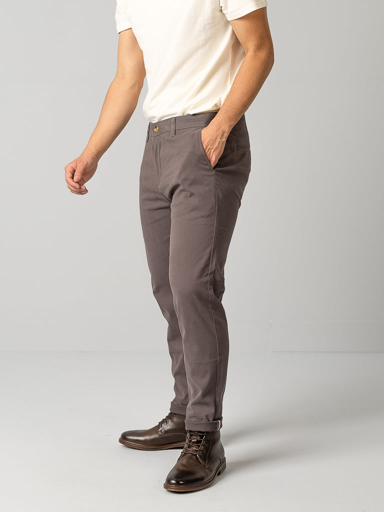 A person in a white t-shirt, gray ID Brooklyn Chino pants from ID Menswear, and brown shoes stands against a plain background. The regular slim fit creates a modern look as they place their left hand in their pocket, slightly turning to the side.
