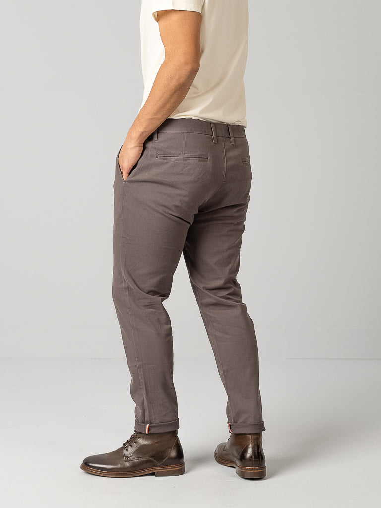 In a minimalist studio, a person poses sideways in cream and gray attire: a t-shirt, ID Brooklyn Chino trousers by ID Menswear made from cotton twill, paired with brown leather ankle boots—a perfect blend of casual elegance.