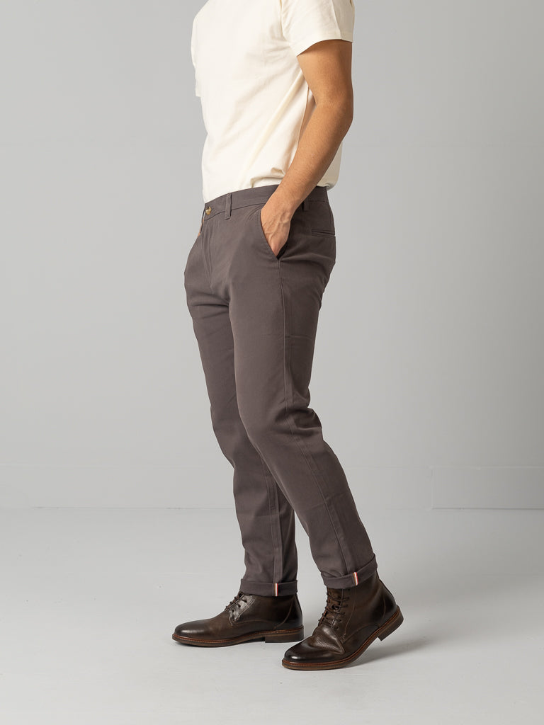 A person in a white t-shirt, gray ID Brooklyn Chino pants from ID Menswear, and dark brown leather boots stands with hands in pockets against a plain background. The casual outfit is neatly presented.