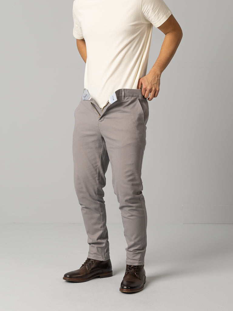 A person in a white t-shirt and gray pants is standing against a plain background. Their dark brown shoes perfectly complement the ID Menswear ID Brooklyn Chino, featuring a regular slim fit with an open button and zipper.