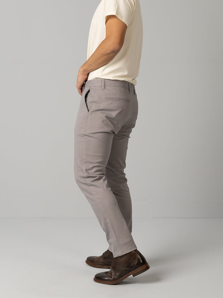 Against a gray backdrop, someone in a cream t-shirt and ID Brooklyn Chino pants by ID Menswear adds chic simplicity to the scene with dark brown leather shoes.