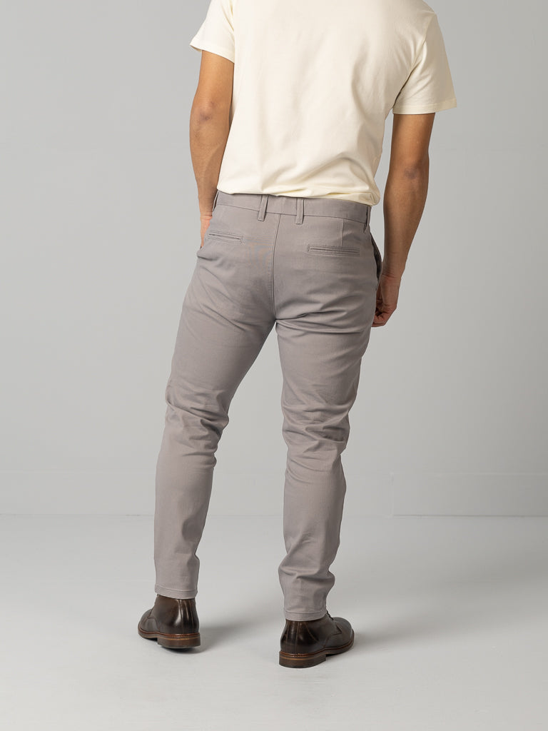 A person stands against a gray backdrop, wearing a white shirt, ID Brooklyn Chino pants in light gray regular slim fit by ID Menswear, and brown shoes. Viewed from behind, their full body is shown dressed in comfortable cotton twill attire.