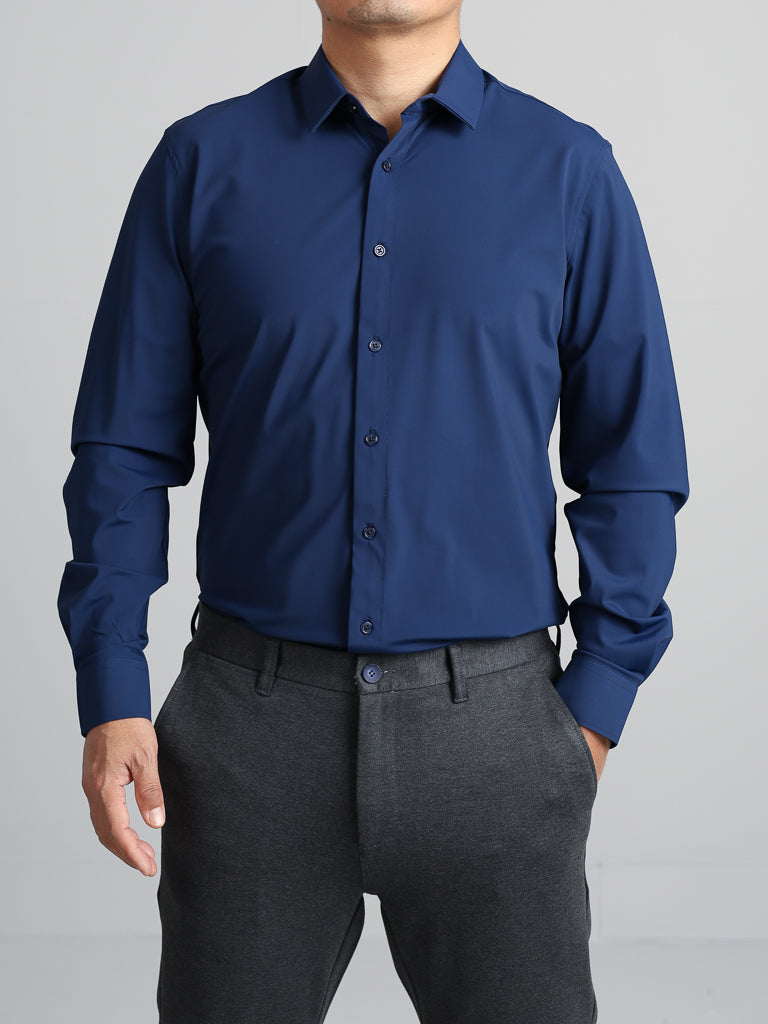 A person models the Axel 4-Way Stretch Dress Shirt by ID Menswear, paired with dark gray trousers. The dark blue slim-fit shirt is designed for comfort and style. The scene features a light gray background, focusing on the upper body with one hand partially in a pocket.