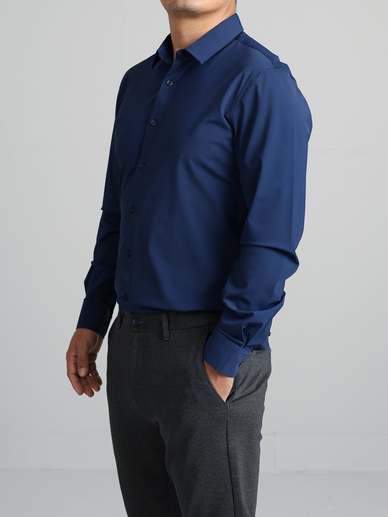 An individual wears the Axel 4-Way Stretch Dress Shirt by ID Menswear, a slim-fit dark blue button-up with a classic collar and long sleeves, paired with gray pants. They pose against a plain light gray background, and their face is not visible.