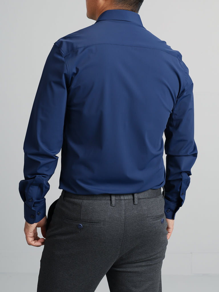 A person is seen from the back in a fitted Axel 4-Way Stretch Dress Shirt by ID Menswear. The dark blue, long-sleeve shirt features buttoned cuffs and pairs with dark gray trousers that have two pockets, combining style and comfort against a plain background.