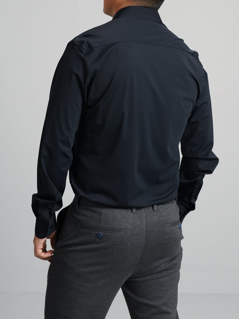 A person is standing with their back to the camera, dressed in comfortable attire—an Axel 4-Way Stretch Dress Shirt from ID Menswear and gray trousers. The 4-way stretch fabric of the shirt complements their silhouette against a plain off-white wall.