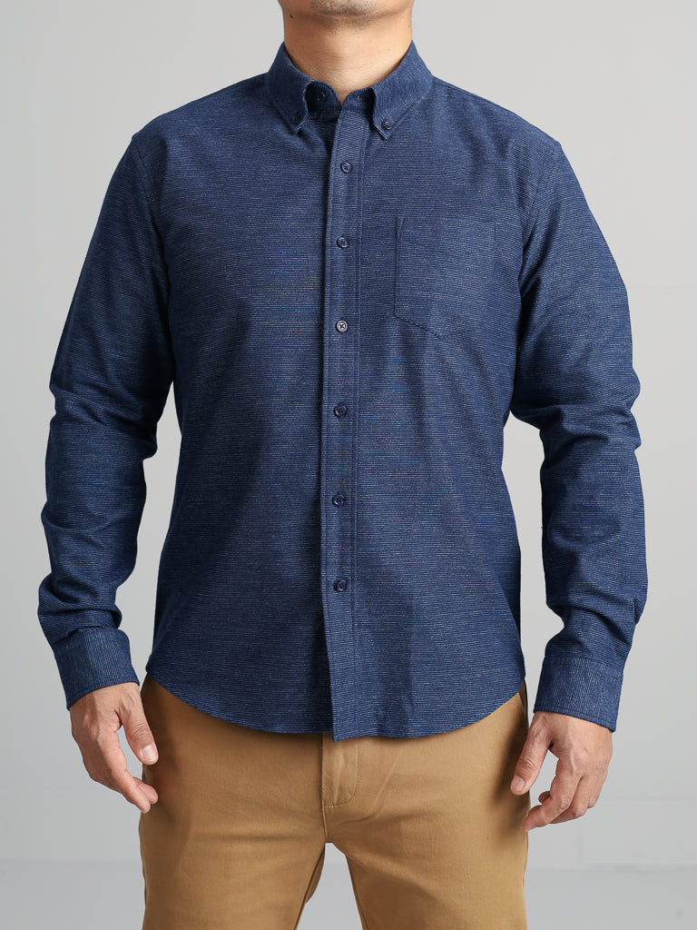 A person wears the Akira Cotton Button Down Shirt by ID Menswear, a dark blue regular-fit shirt with long sleeves, a button-down collar, and a left-side pocket. Paired with beige pants against a light gray background, the image focuses on their torso.