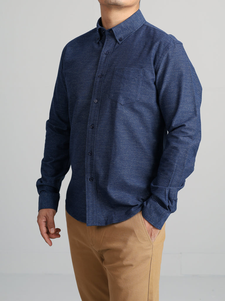 A person stands against a light gray background wearing the Akira Cotton Button Down Shirt by ID Menswear, a dark blue regular-fit long sleeve shirt with a button-down collar and chest pocket, paired with khaki pants. The persons head is not visible.