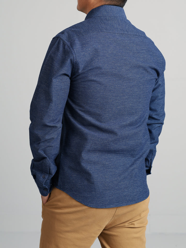 A person wearing an Akira Cotton Button Down Shirt by ID Menswear in blue, regular-fit, textured fabric with a button-down collar and khaki pants is facing away from the camera, with their hands resting casually at their sides.