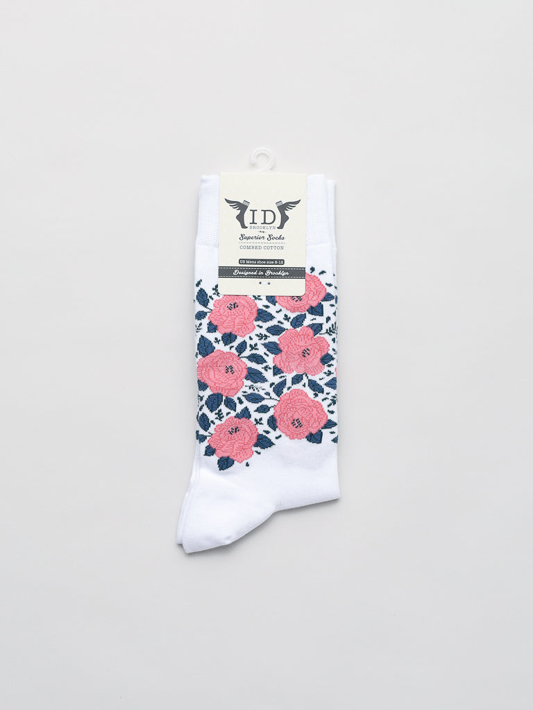 The Roses Graphic Socks from ID Menswear are white, featuring a floral pattern of pink roses and green leaves, made from premium combed cotton. Neatly folded with a cream-colored label showcasing a winged logo and text, they sit against a plain light gray background to enhance their elegance.