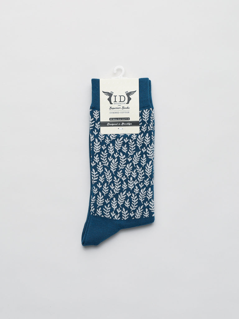 The Flora Graphic Socks by ID Menswear, showcasing a blue design with a white leaf pattern, are elegantly displayed on a plain backdrop. A cardboard tag labeled Flora Graphic Socks is attached, hanging from a white hook at the top, promising premium comfort and style.