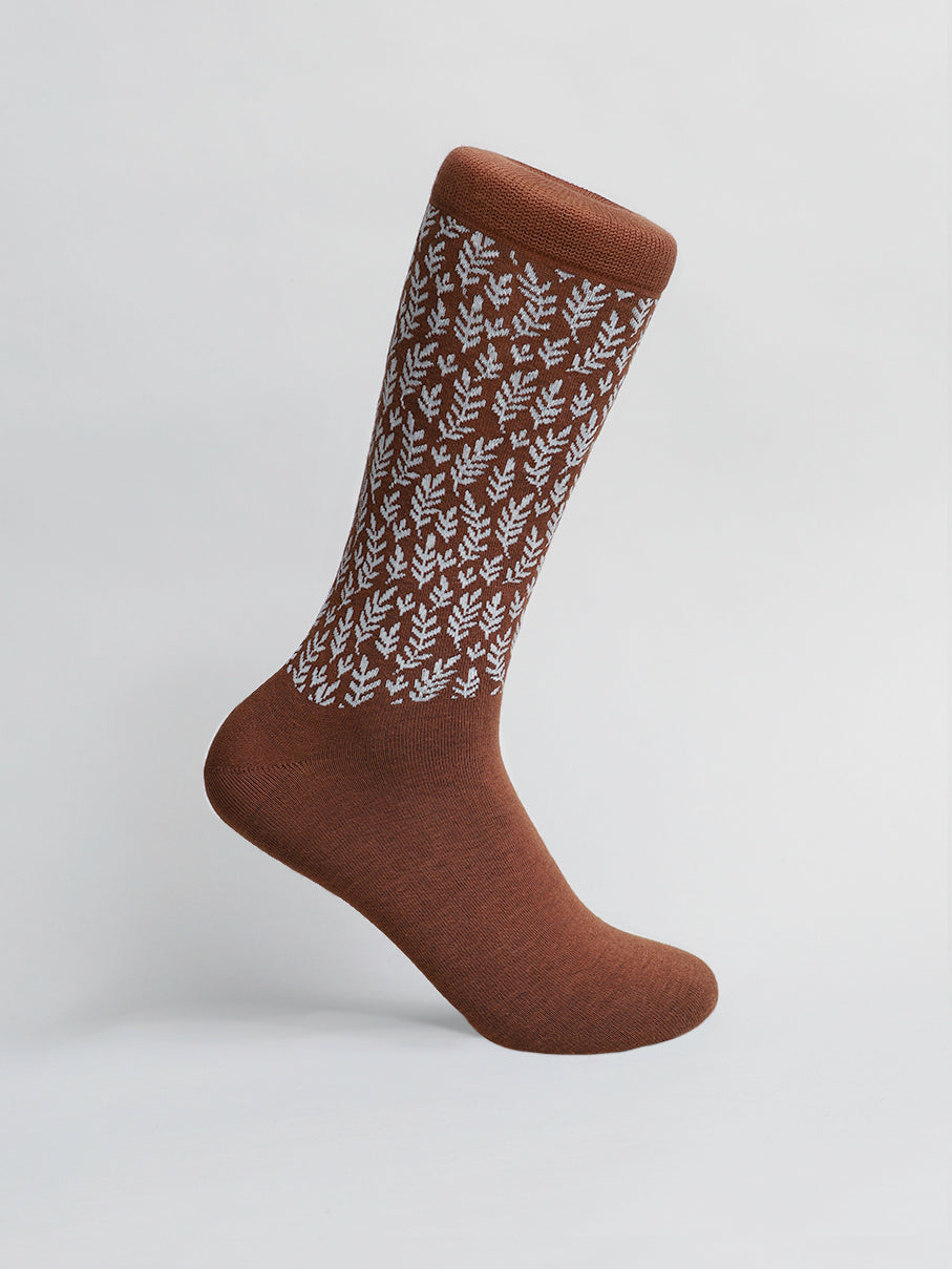 A single Flora Graphic Sock by ID Menswear, featuring a brown color with a white leaf pattern, is shown against a light gray background. It stands upright, showcasing its premium design and texture.