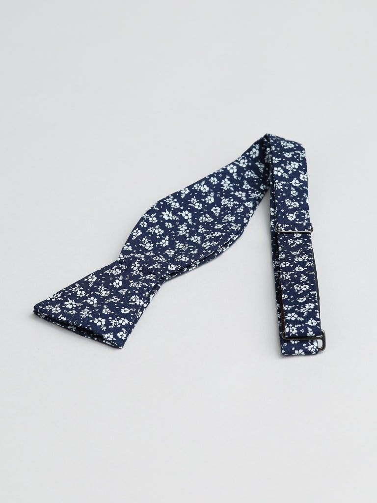 The Ditsy Cotton Bow Tie by ID Menswear features a navy blue and white floral pattern, handcrafted in Brooklyn from cotton. Shown flat on a light gray background, it’s an ideal semi-formal accessory.