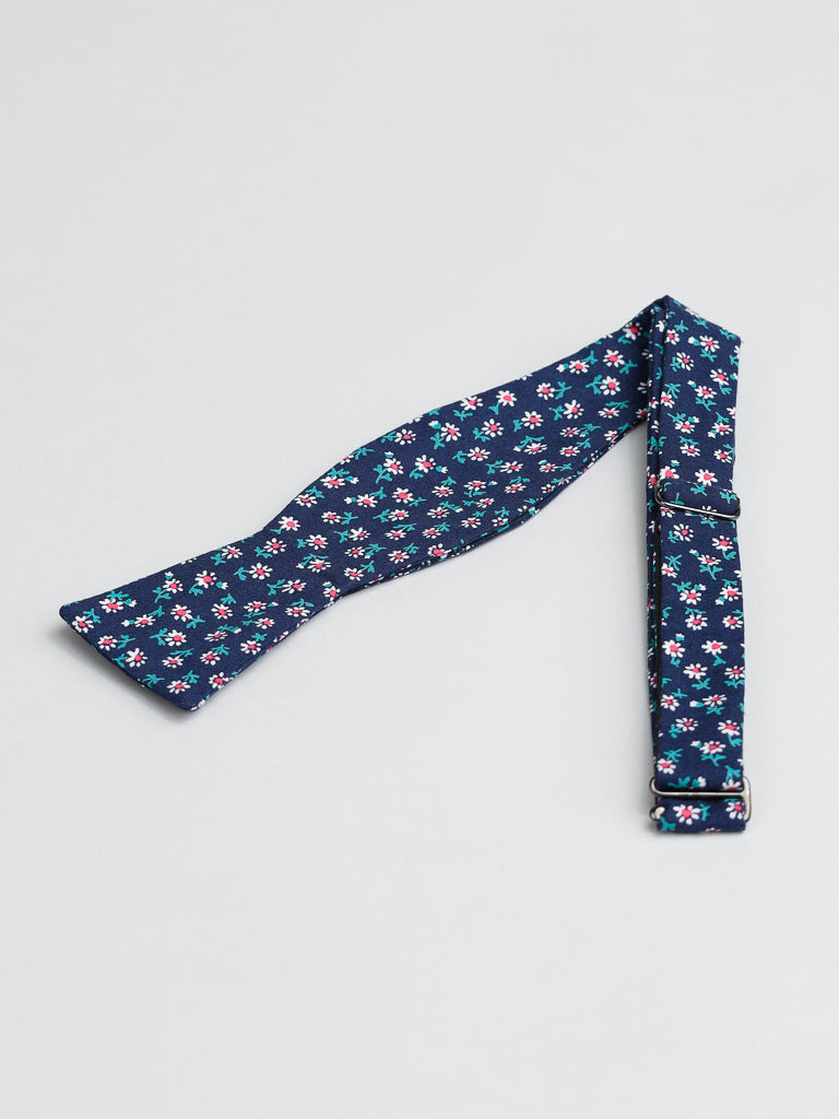 The White Bloom Cotton Bow Tie by ID Menswear is a navy blue, 100% cotton accessory with small white and pink flowers. Handmade in Brooklyn, it’s untied and includes an adjustable strap with metal clasp. Shown flat on a light gray surface.