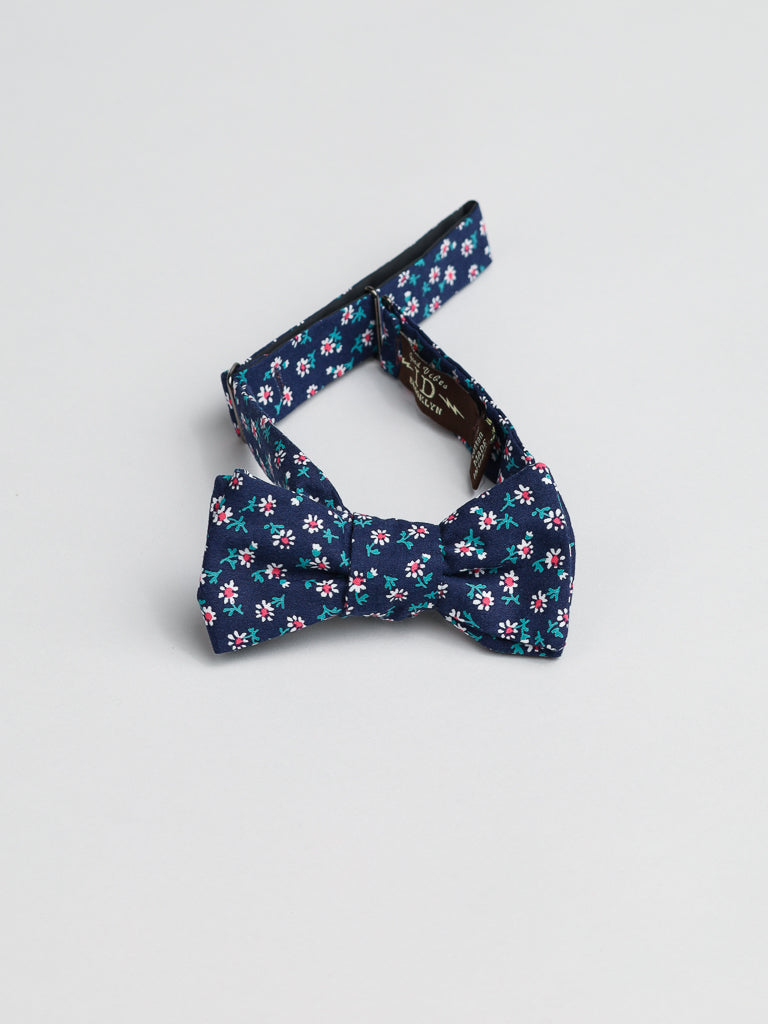 The White Bloom Cotton Bow Tie by ID Menswear features small white, pink, and teal flowers on navy blue cotton. Handmade in Brooklyn, it has adjustable straps extending from each side and is displayed on a light gray background.