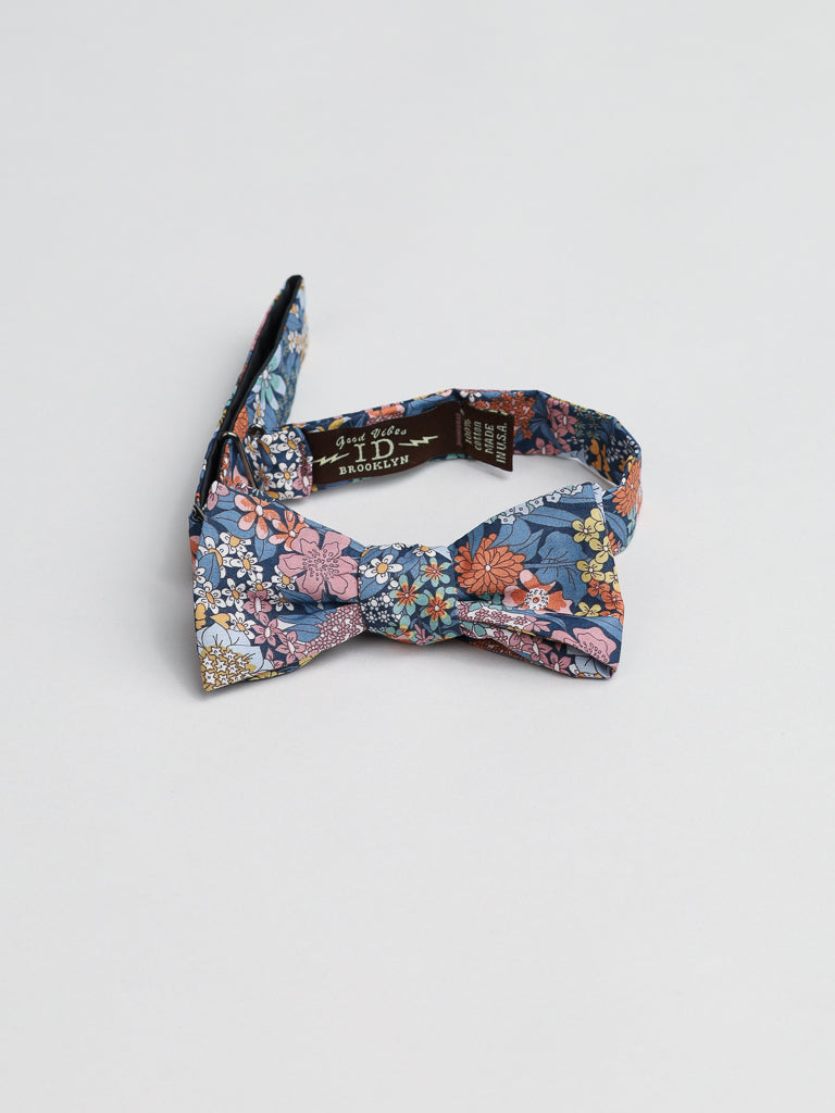 The ID Menswear Liberty of London Tana Lawn™ Ciara Cotton Bowtie features a floral design with blue, pink, orange, and yellow hues on white. Handmade in Brooklyn, its placed on light gray fabric.