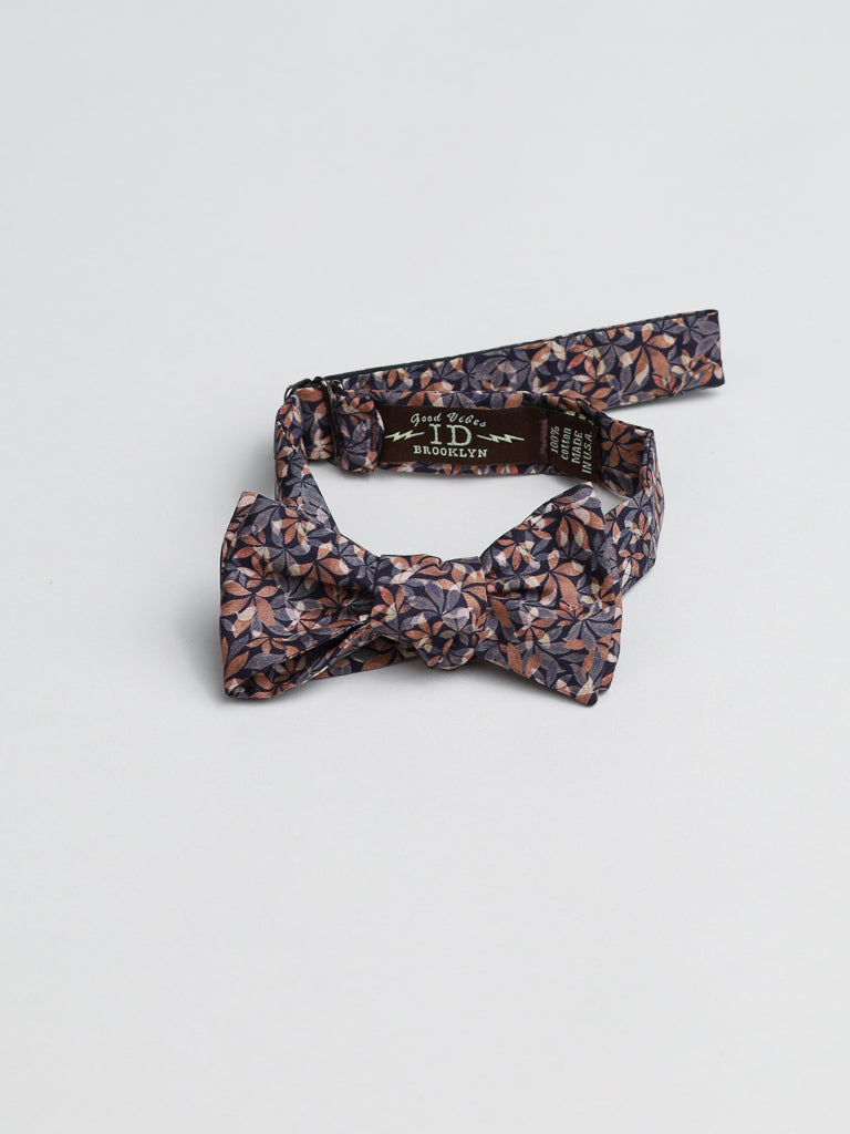 A floral-patterned ID Liberty of London Tana Lawn™ Leaves Cotton Bowtie from ID Menswear, featuring a dark background and adjustable strap, lies on a light gray surface. The inside tag partially shows handmade in Brooklyn in crisp white text.