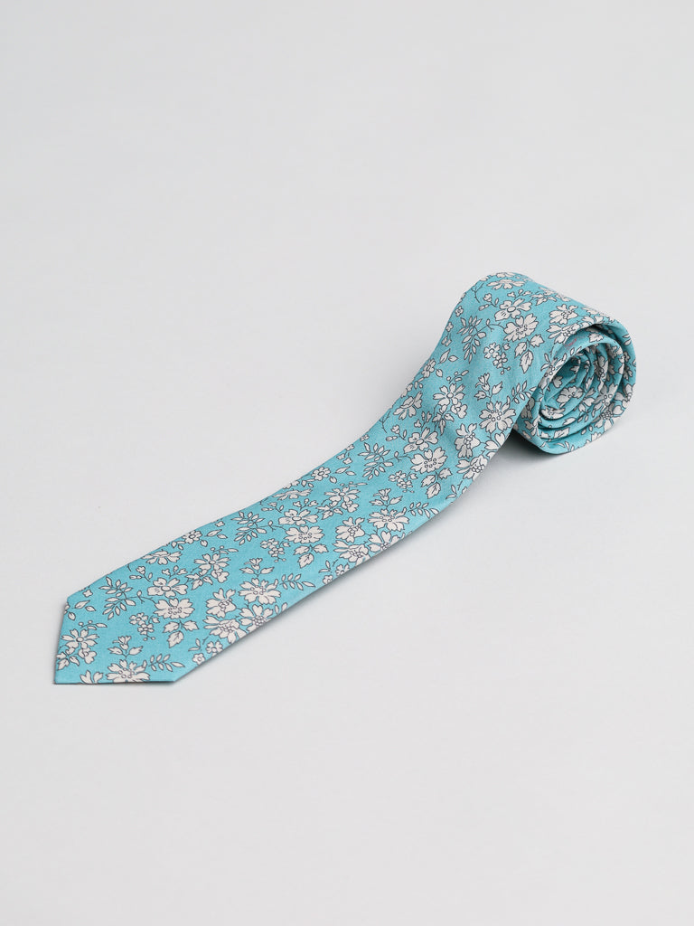 A Liberty of London Tana Lawn™ Carpel Tie by ID Menswear, handmade in Brooklyn from light blue cotton with a white and gray floral pattern, is shown rolled up on a plain white background.
