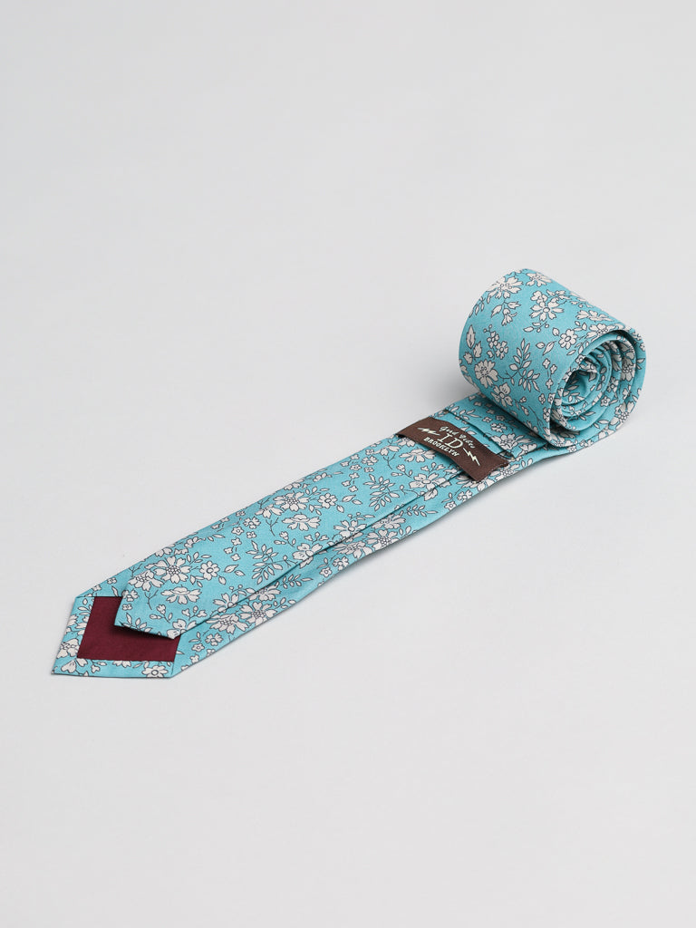 A rolled-up Liberty of London Tana Lawn™ Carpel Tie by ID Menswear, light blue with a white floral pattern and maroon inner tip, handmade in Brooklyn and displayed on a plain light background.
