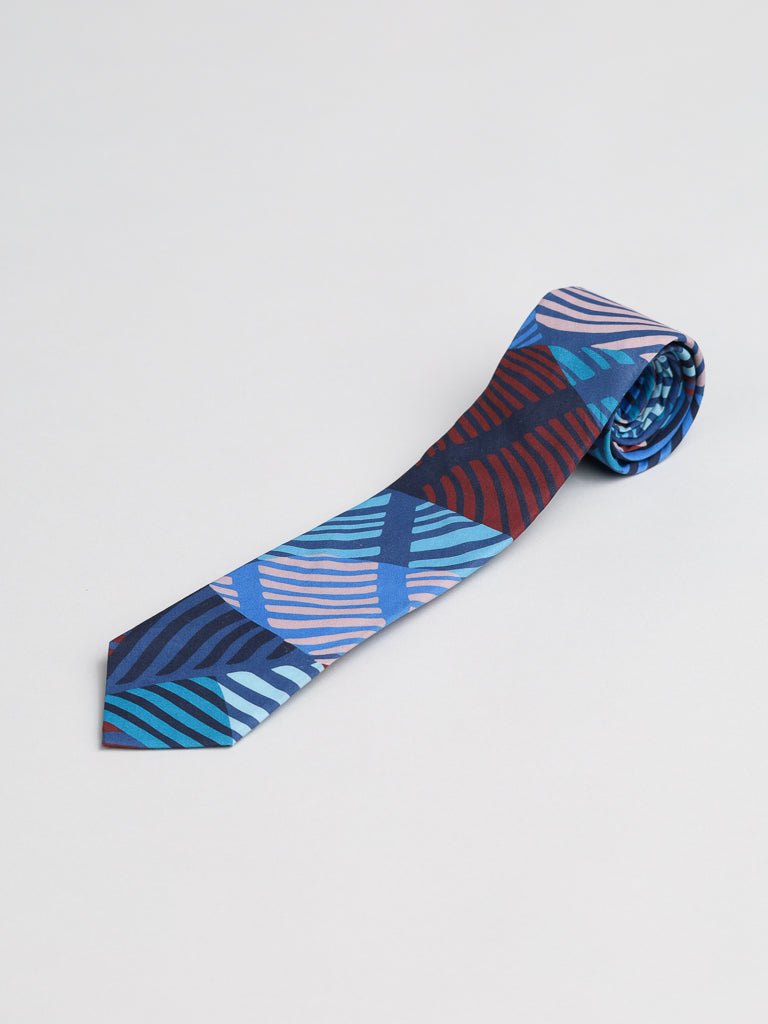 The Liberty of London Tana Lawn™ Wilde Tie by ID Menswear features bold abstract leaves in blue, teal, purple, and red on dark cotton, handmade in Brooklyn and shown rolled up on a light surface.