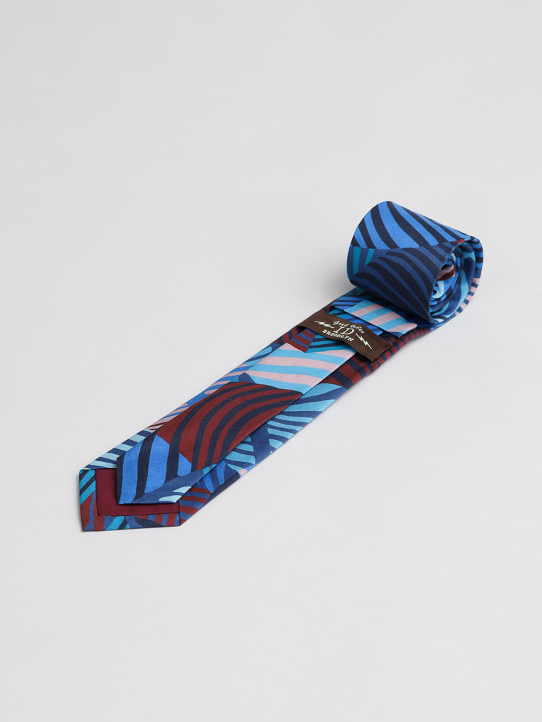 A rolled-up Liberty of London Tana Lawn™ Wilde Tie by ID Menswear, handmade in Brooklyn with a bold abstract blue, red, and light purple pattern, displayed on a plain white background.
