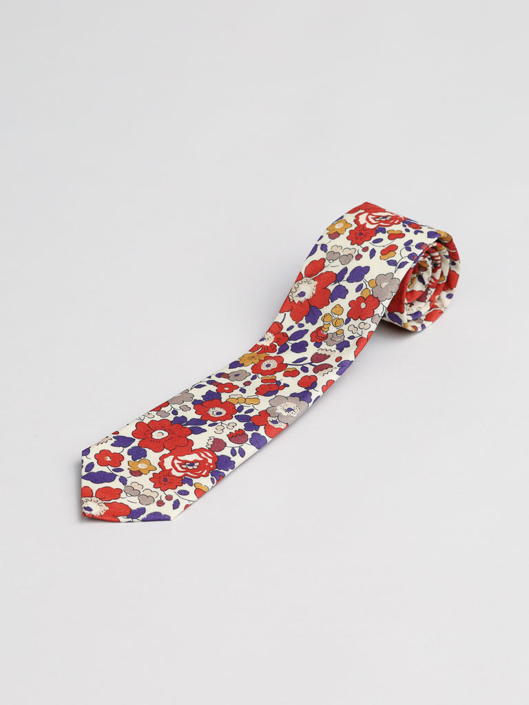 A Liberty of London Tana Lawn™ Betsy Tie by ID Menswear, handmade in Brooklyn from soft cotton, featuring a vibrant floral pattern with red, orange, and purple flowers on white, is rolled up and displayed on a plain white surface.