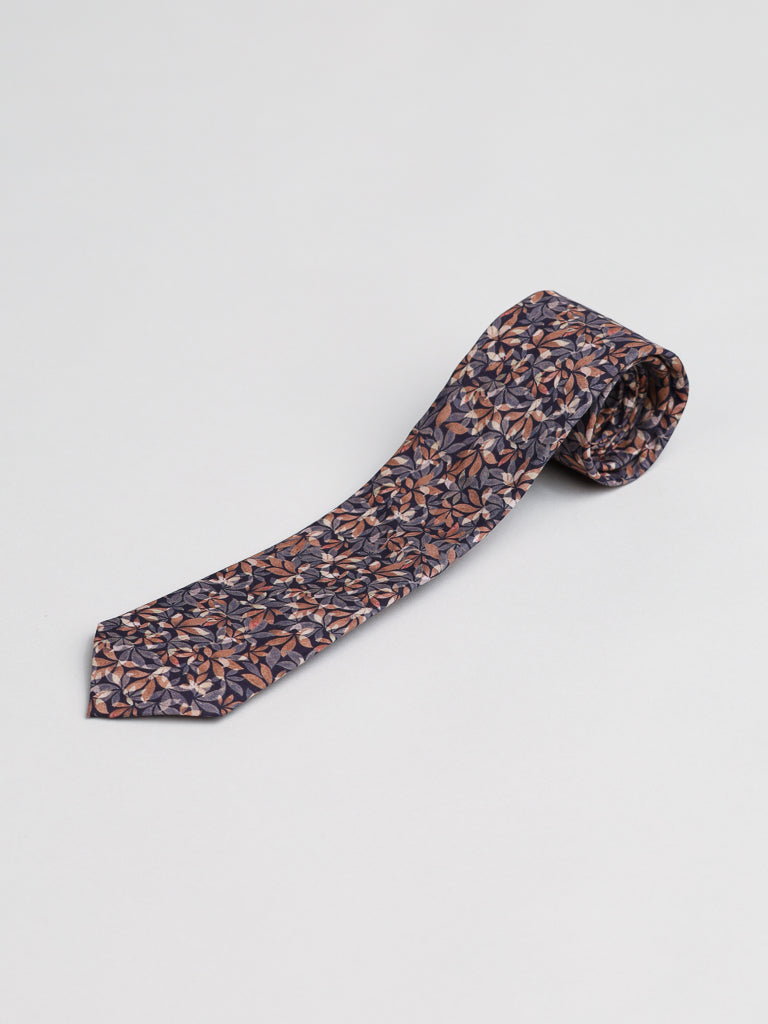 The Liberty of London Tana Lawn™ Leaves Tie by ID Menswear features a dark silk background with an all-over multicolored leaf pattern in brown, tan, and blue. Handmade in Brooklyn, it’s displayed on a light gray surface.