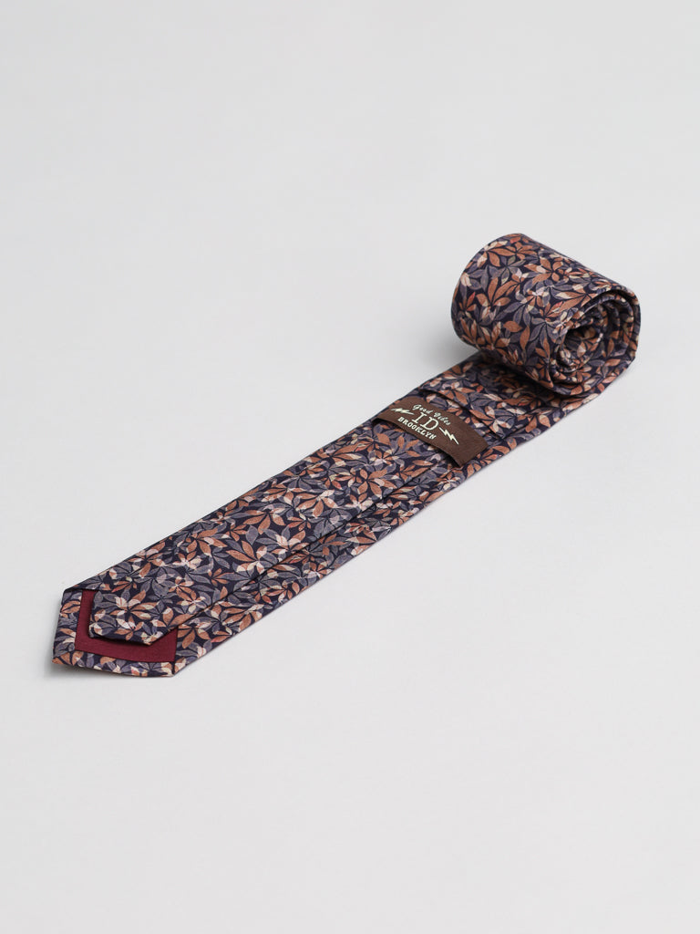 The Liberty of London Tana Lawn™ Leaves Tie by ID Menswear, handmade in Brooklyn, features a multicolored brown, blue, and beige leaf pattern with a silk-like feel, shown rolled up diagonally on a plain white surface.
