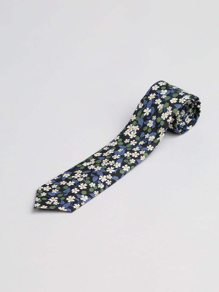 The Liberty of London Tana Lawn™ Karen Tie by ID Menswear, handmade in Brooklyn from luxurious cotton, has a navy base with white, green, and blue floral patterns, shown rolled up on a light gray surface.