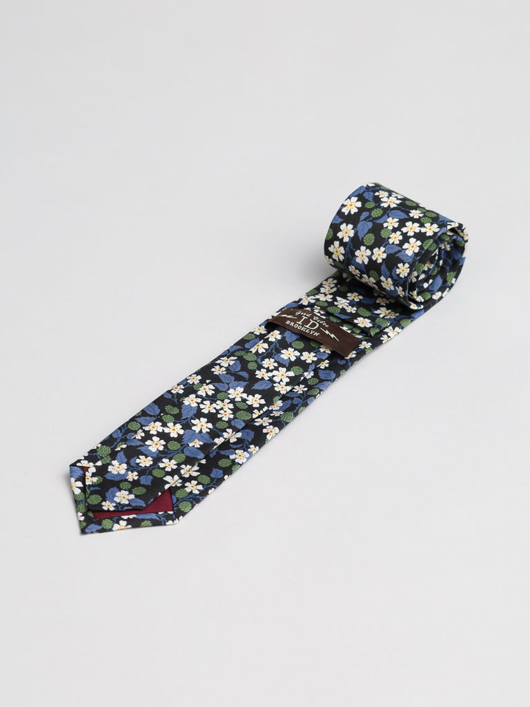 The Liberty of London Tana Lawn™ Karen Tie by ID Menswear is a navy blue tie with small white and yellow flowers and green leaves, crafted from Tana Lawn cotton, shown rolled up on a light grey background.