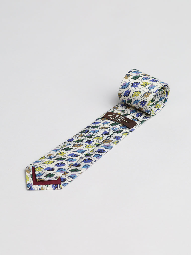 The ID Menswear Liberty of London Tana Lawn™ Sunrise Tie, handmade in Brooklyn, displays blue, green, and yellow abstract shapes on premium cotton. One end is rolled while the tie rests on a plain white surface.